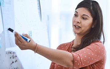 Dark-haired woman wearing a mauve polkadot shirt, holding an Expo marker training a defense support team