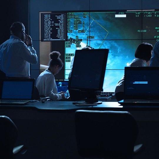 Two women and a man wearing a headset looking at a large screen in a control room